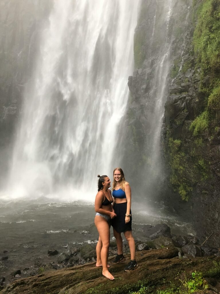 Two women standing by a waterfall in Tanzania, embracing nature and adventure.